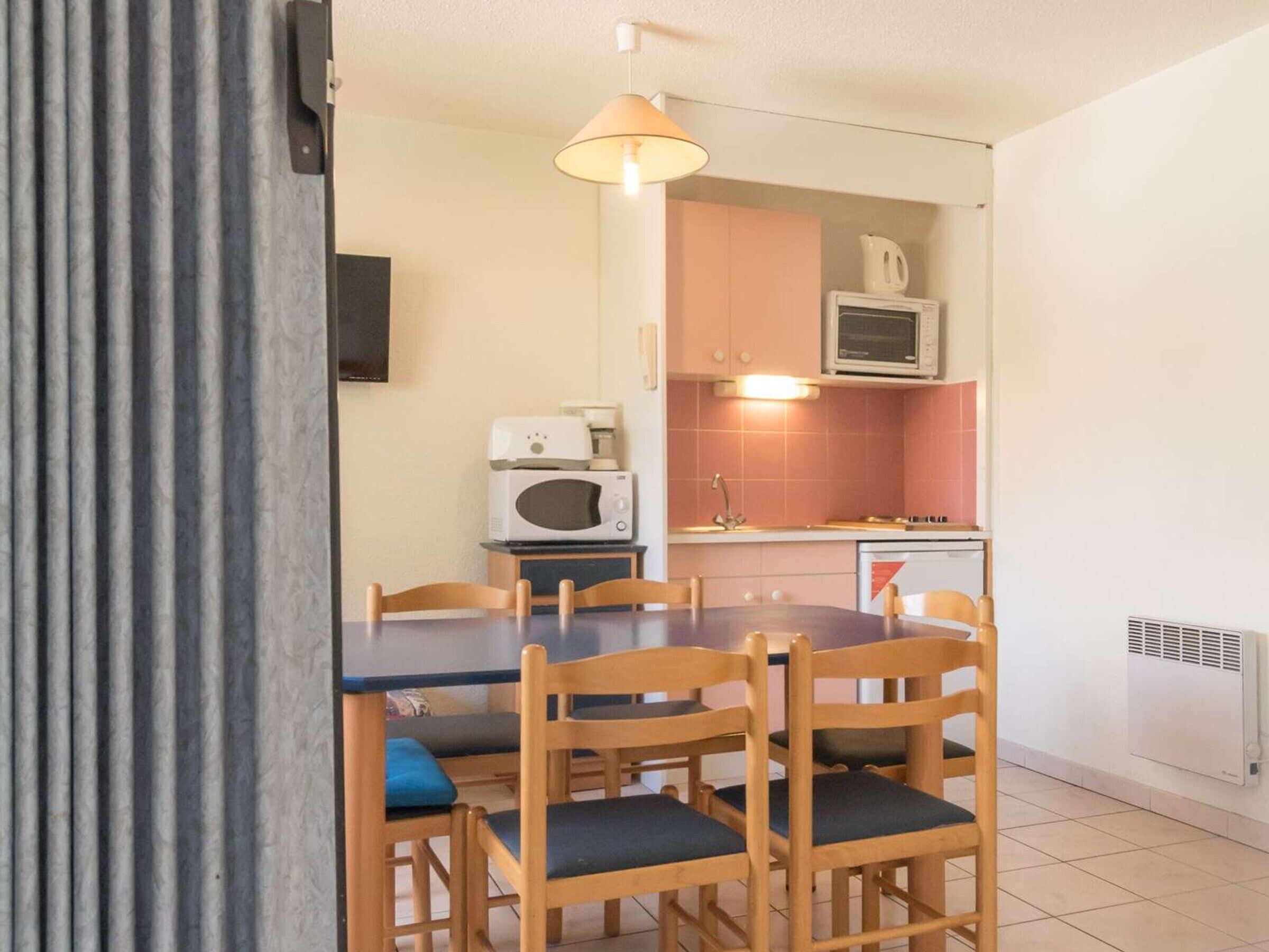 A compact kitchen with a microwave, coffee maker, and a small sink. There is a dining table with four chairs, and a ceiling light fixture above.