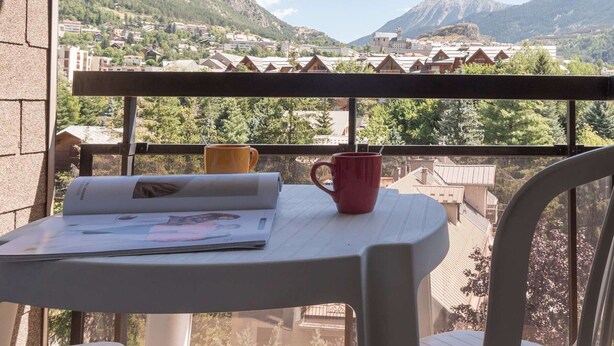 A balcony with a table, a red mug, and a newspaper. The view includes a mountain and a village.