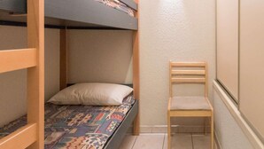 A bunk bed with a patterned bedspread, a pillow, and a wooden chair in a narrow room with a textured wall and a ceiling light.