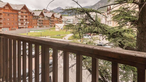 A view of a hotel with multiple buildings, including a prominent red building with balconies. There is a tree in the foreground, and the background features mountains.