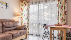 A cozy living room with a brown sofa, a wooden table, and a chair. There is a window with sheer curtains and a colorful leaf-patterned curtain.