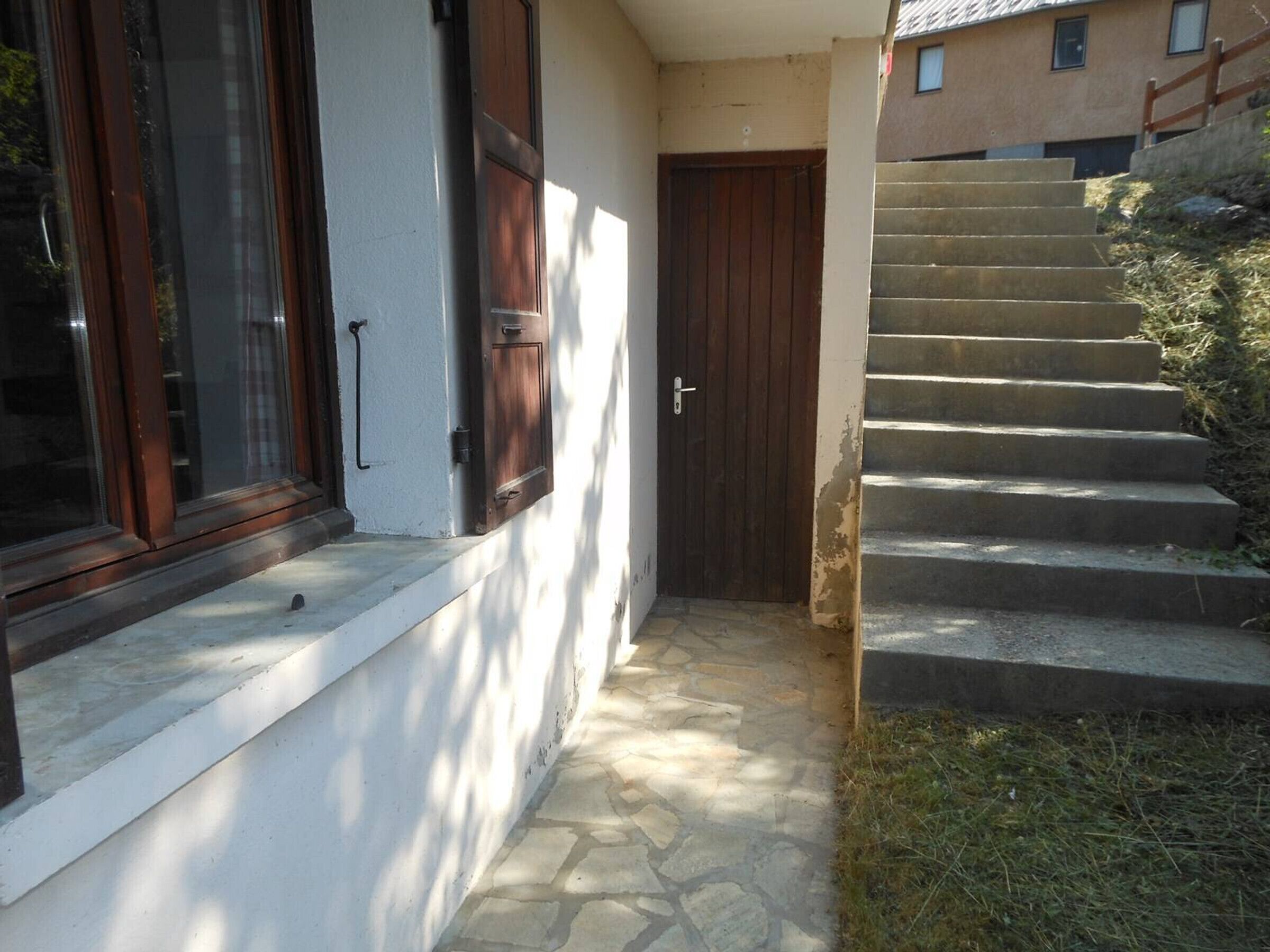Un étroit sentier de pierre mène à une porte en bois, flanquée d’un mur blanc et d’une fenêtre avec des volets en bois.