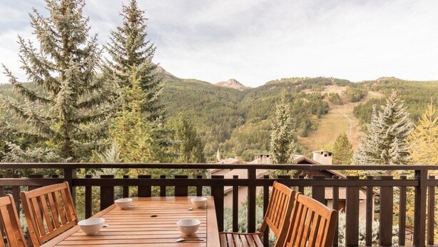 Une table en bois, quatre chaises et des bols sont disposés sur un balcon qui offre une vue sur un paysage montagneux.