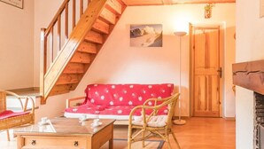 Une pièce chaleureuse avec un escalier en bois, un canapé rouge orné d’un motif en forme de flocon de neige, un fauteuil en osier, une table en bois avec des tasses, et un tableau accroché au mur.
