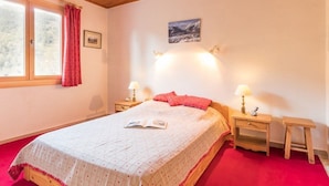 Une chambre avec un plafond en bois, un lit recouvert d’une courtepointe rouge et blanche, deux tables de chevet avec des lampes, une fenêtre avec des rideaux rouges et un tableau encadré au mur.
