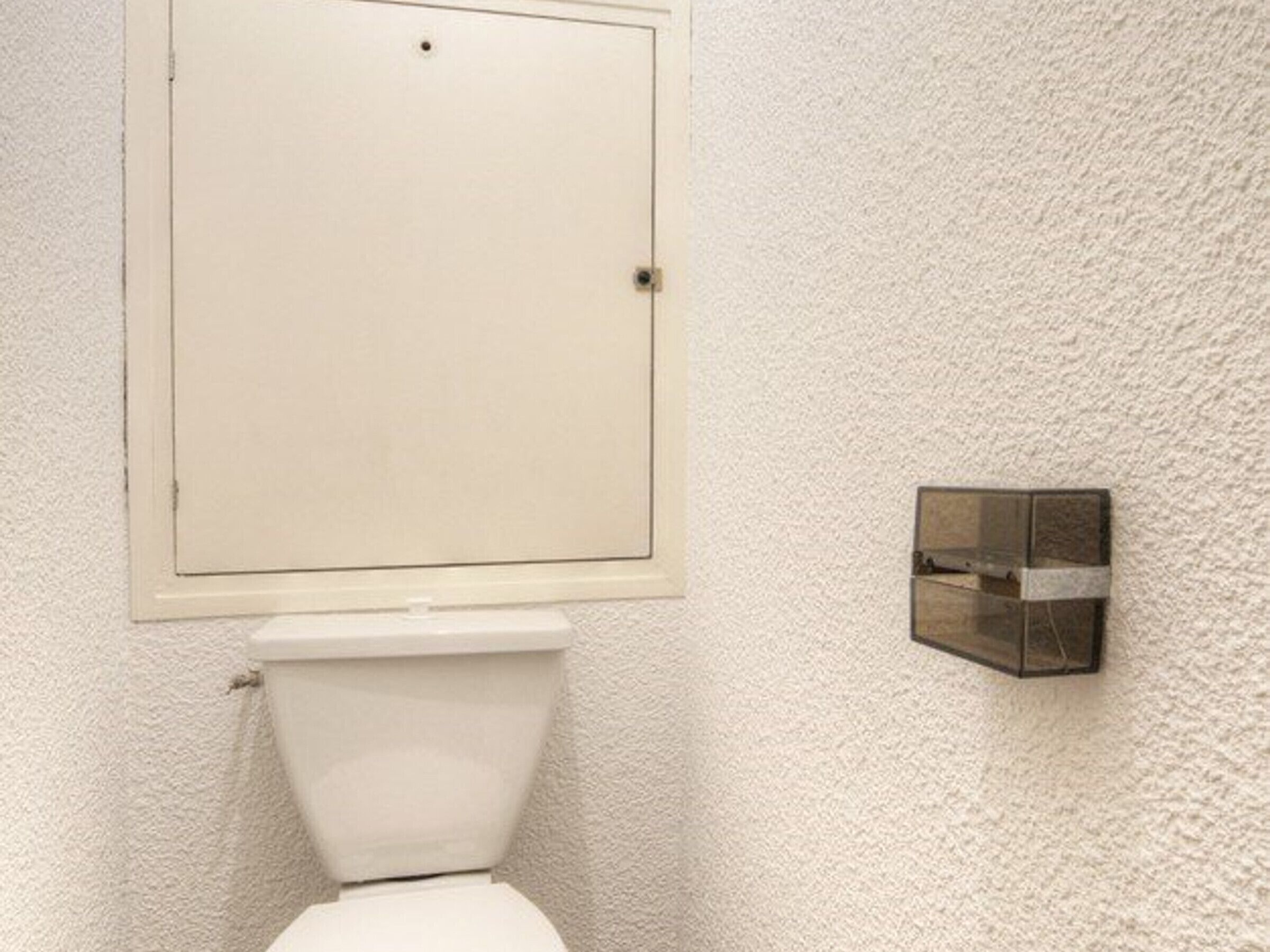 Ein Badezimmer mit einer weißen Toilette, einer strukturierten weißen Wand und einem kleinen, dunkel gefärbten Schrank.