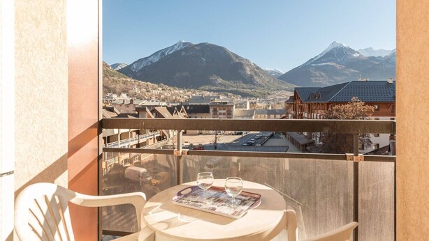 A balcony with a table and chairs, overlooking a mountain range and a town.