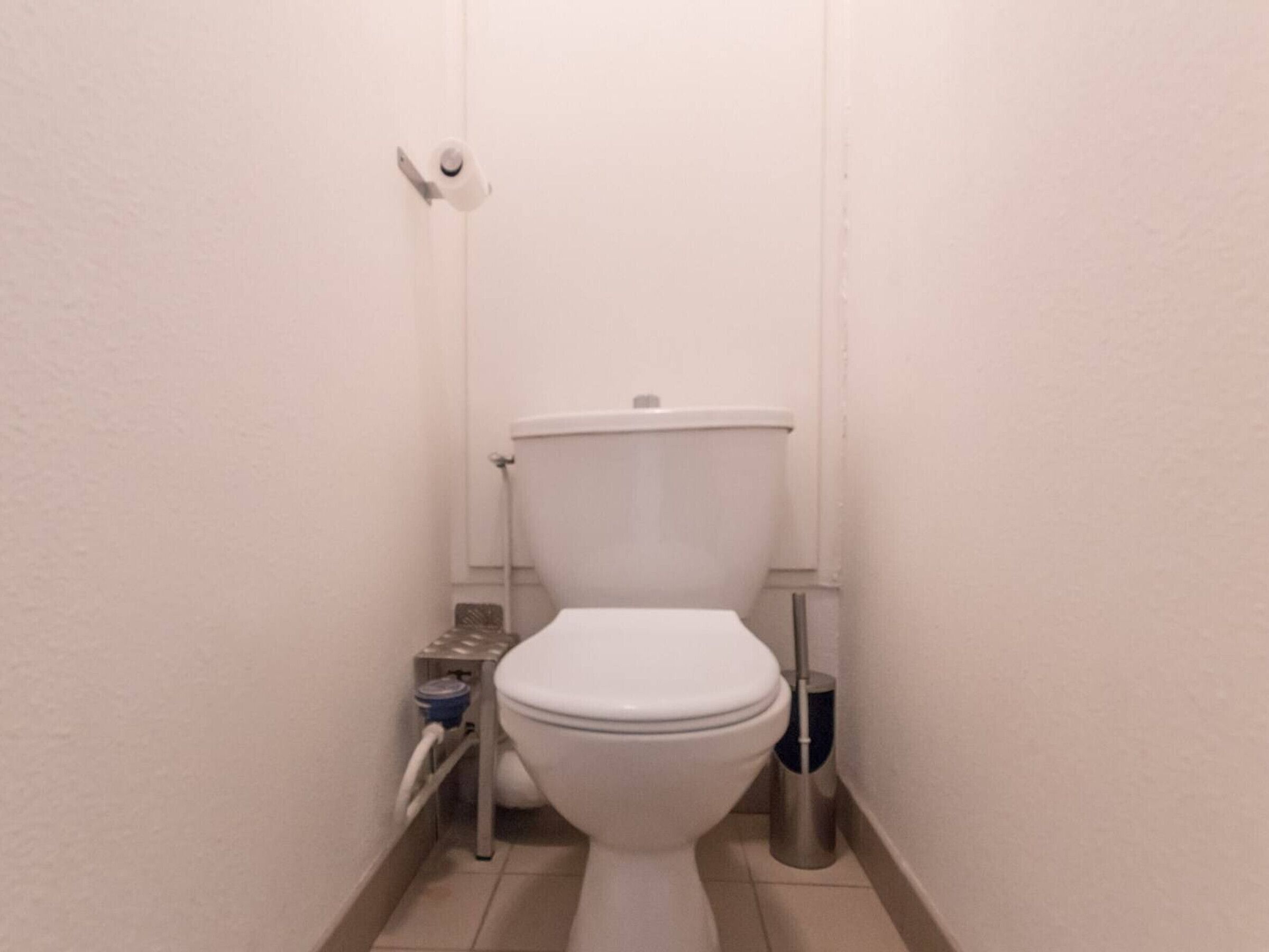 Een klein, wit toilet in een smalle badkamer met een witte muur en betegelde vloer.