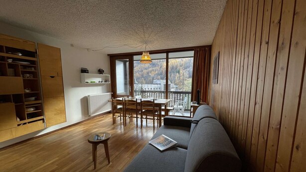 Een moderne woonkamer met houten vloer, een grijze bank, een eettafel met stoelen, een klein bijzettafeltje en een groot raam met uitzicht op bomen en een gebouw.