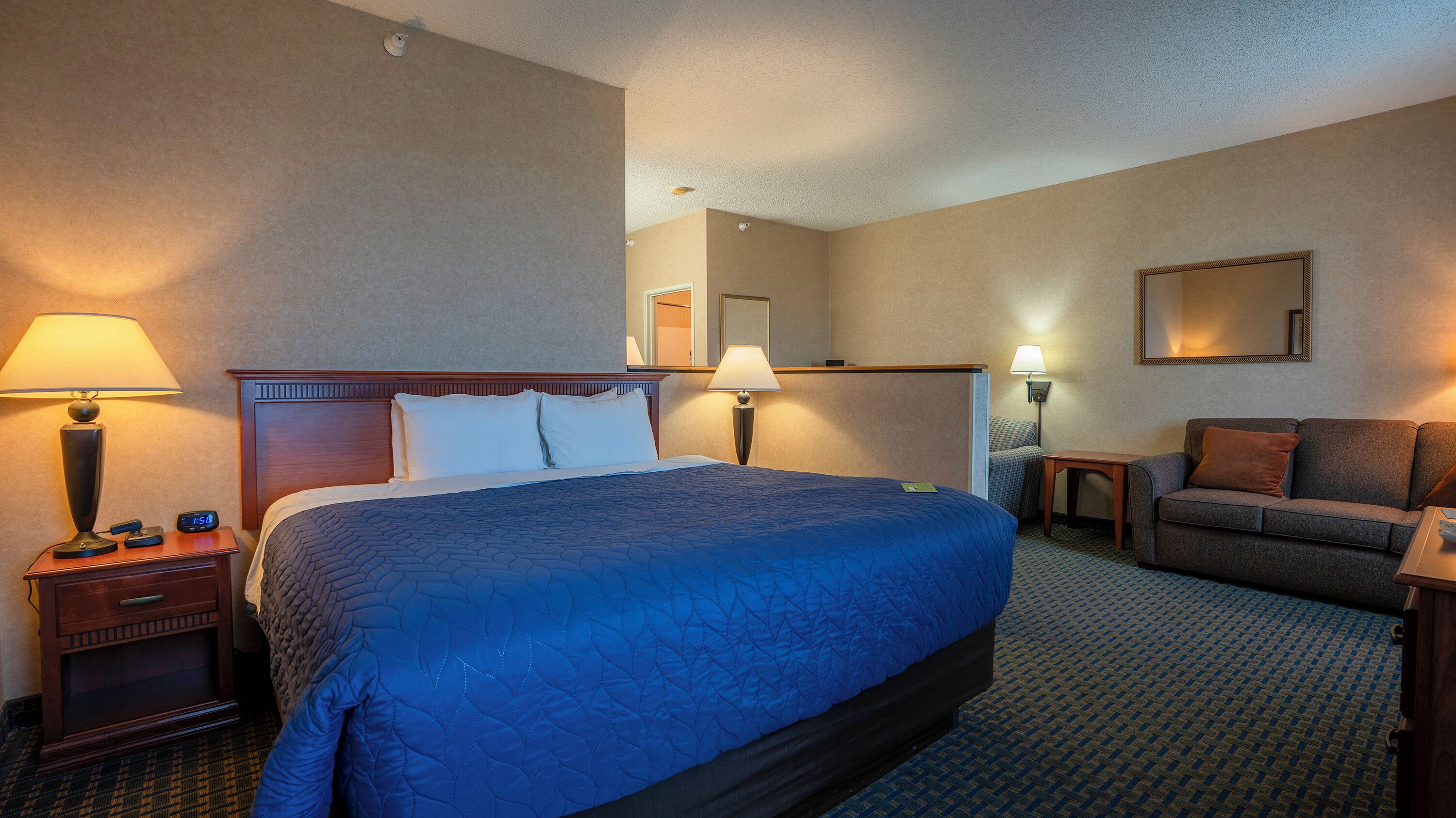 A hotel room with a large bed, bedside tables with lamps, a small table, a sofa, and a mirror on the wall.
