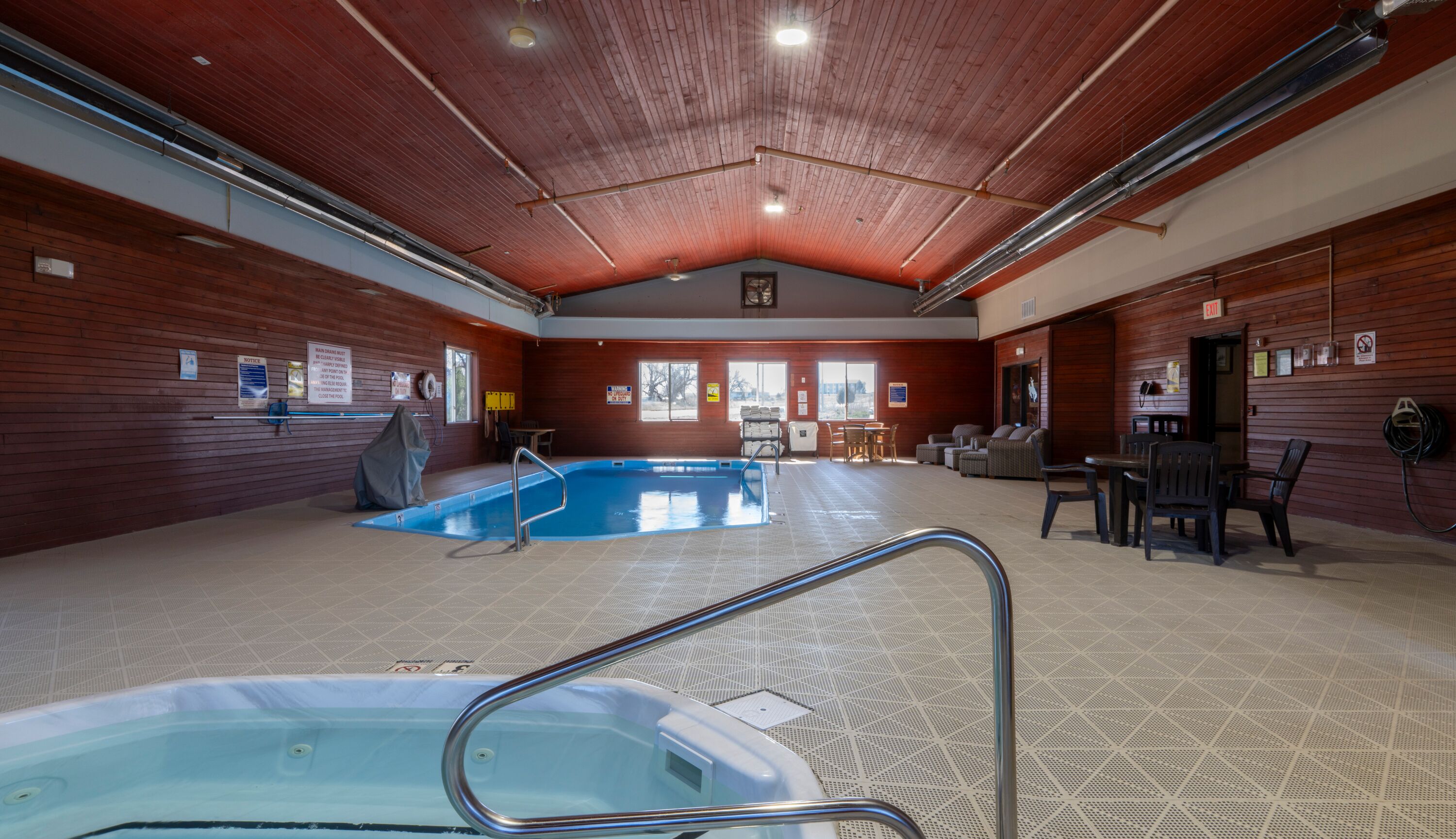 Una piscina interior con un jacuzzi, paredes de madera y una zona de estar con mesas y sillas.