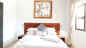Una cama bien hecha con sábanas blancas, cabecera de madera y una obra de arte enmarcada encima.