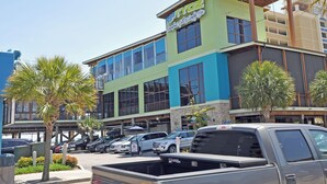 A multi-story building with a green and blue facade, large windows, and a parking lot with several cars. - 3BR Condo in Myrtle Beach: Perfect for Families (MYRTLE BEACH)