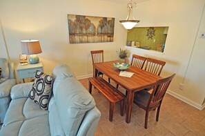 A dining area with a wooden table, chairs, a bench, a lamp, a painting, and a small plant on a shelf. - 3BR Condo in Myrtle Beach: Perfect for Families (MYRTLE BEACH)
