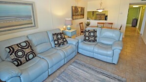 A living room with two blue leather sofas, patterned throw pillows, a small side table, a painting, and a ceiling fan. - 3BR Condo in Myrtle Beach: Perfect for Families (MYRTLE BEACH)