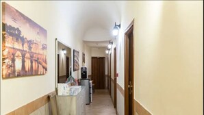 A hallway with a reception desk, a chair, a refrigerator, and a door at the end. - Alex Romano (Rome)