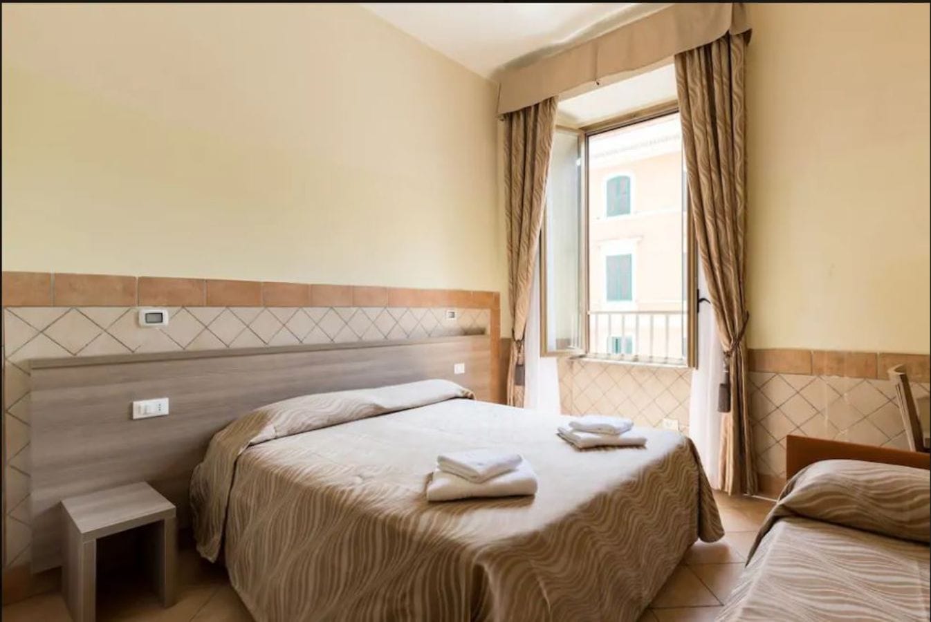 A hotel room with a bed, a bedside table, and a window with curtains.