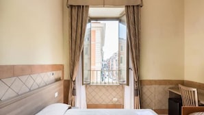 A hotel room with a bed, a desk, and a view of buildings through a window with curtains. - Alex Romano (Rome)