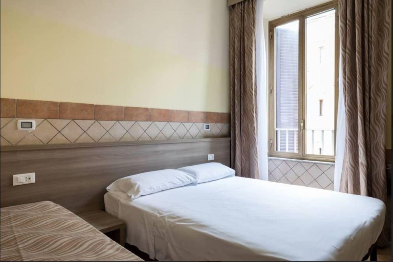 Habitación de hotel con cama, mesita de noche, toma de corriente y ventana con cortinas.