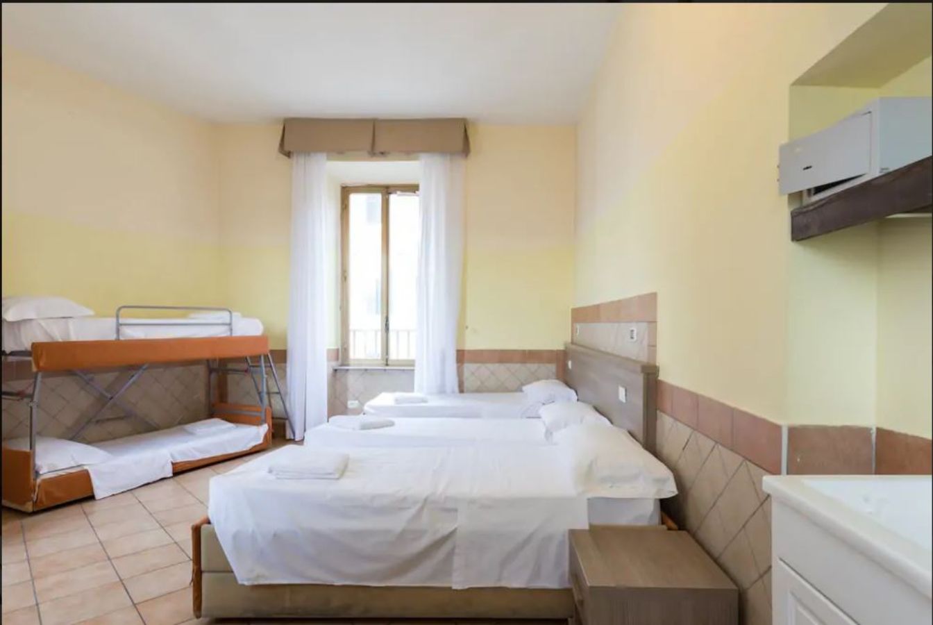 A hotel room with two beds, a window with curtains, a built-in air conditioner, and a small bedside table.
