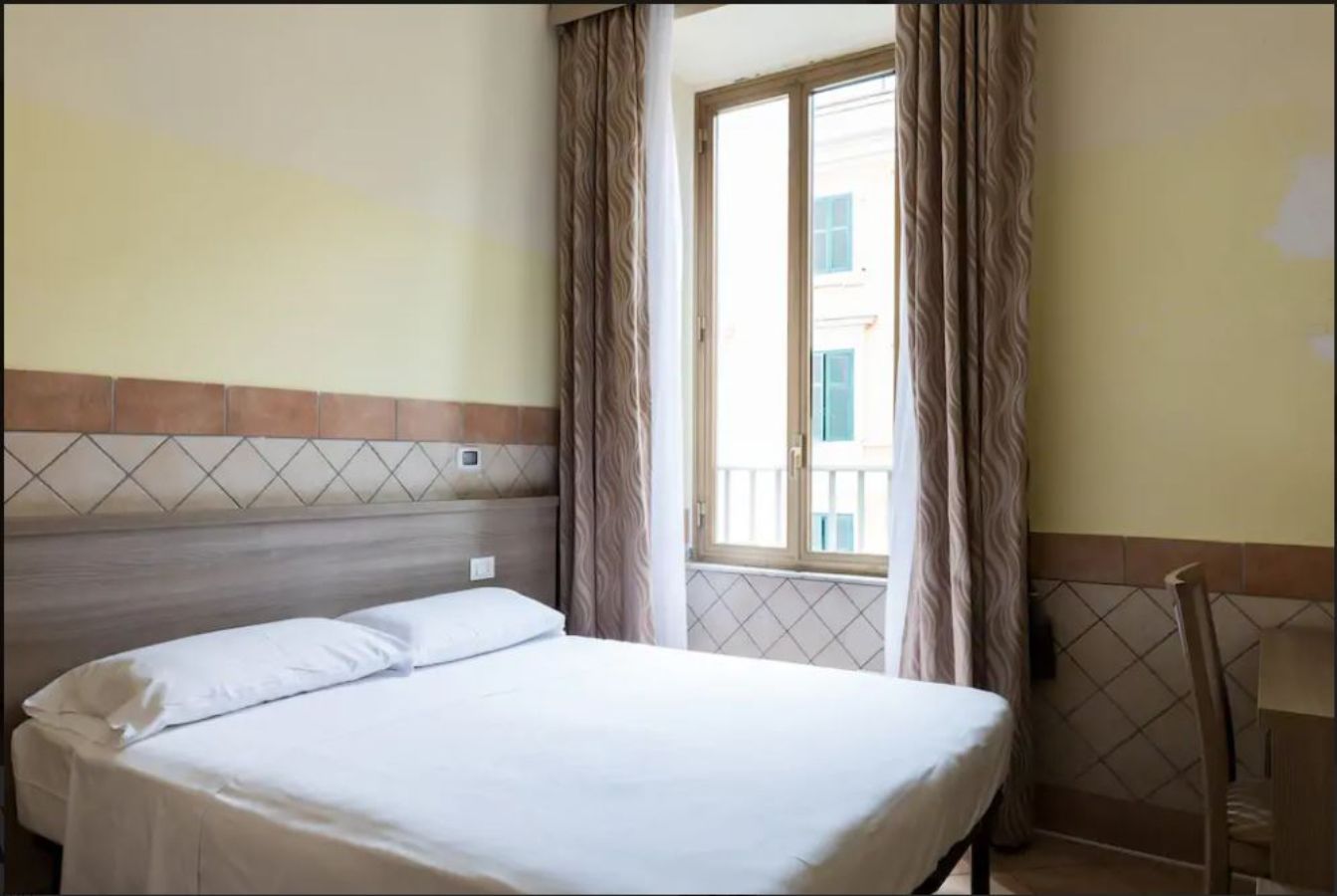Quarto de hotel com cama, janela com cortinas e vista para um edifício.
