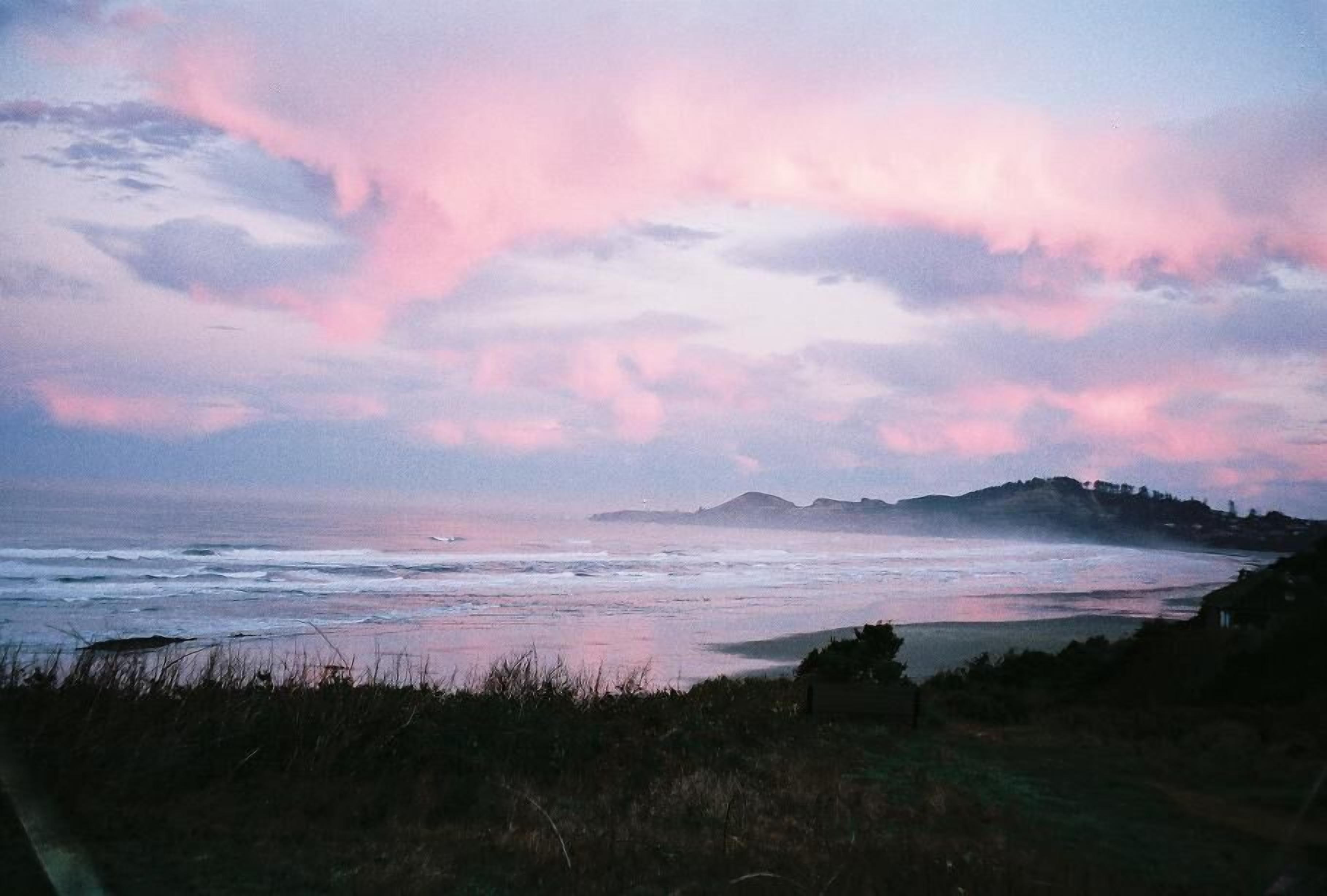 Een kustlandschap met een strand, oceaan en heuvels in de verte onder een roze en blauwe lucht.