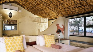 A hotel room with a thatched roof, a bed with patterned pillows, a wall-mounted air conditioner, and a window with a view of trees. - Ugab Terrace Lodge (Khorixas)
