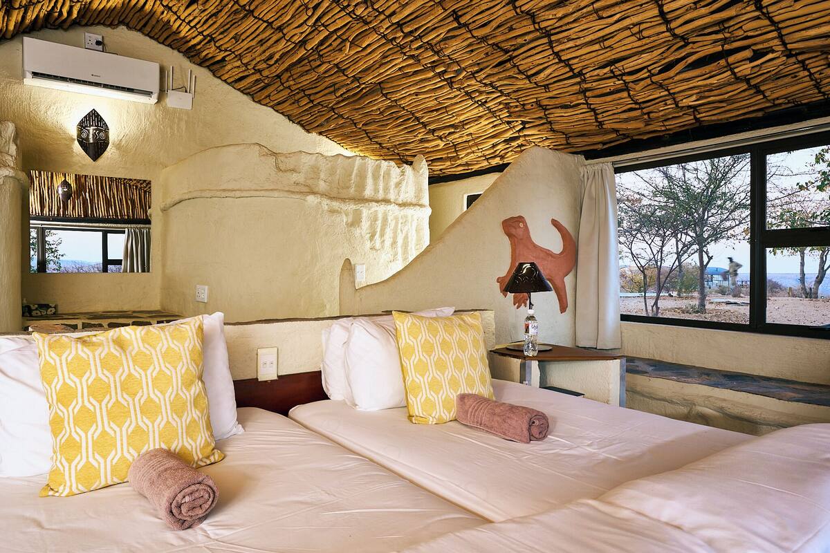 A hotel room with a thatched roof, a bed with patterned pillows, a wall-mounted air conditioner, and a window with a view of trees.