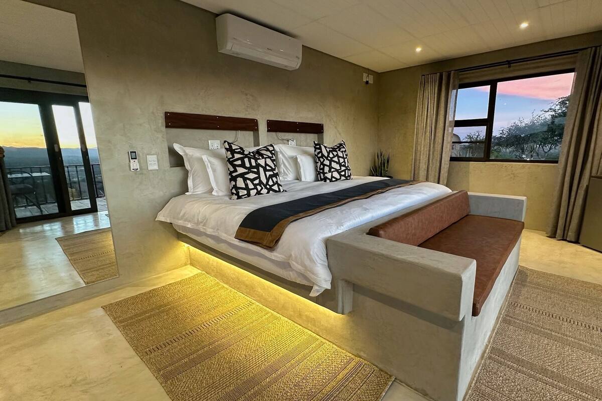 A modern bedroom with a large bed, a bedside table, a built-in bench, and a view of the outdoors.