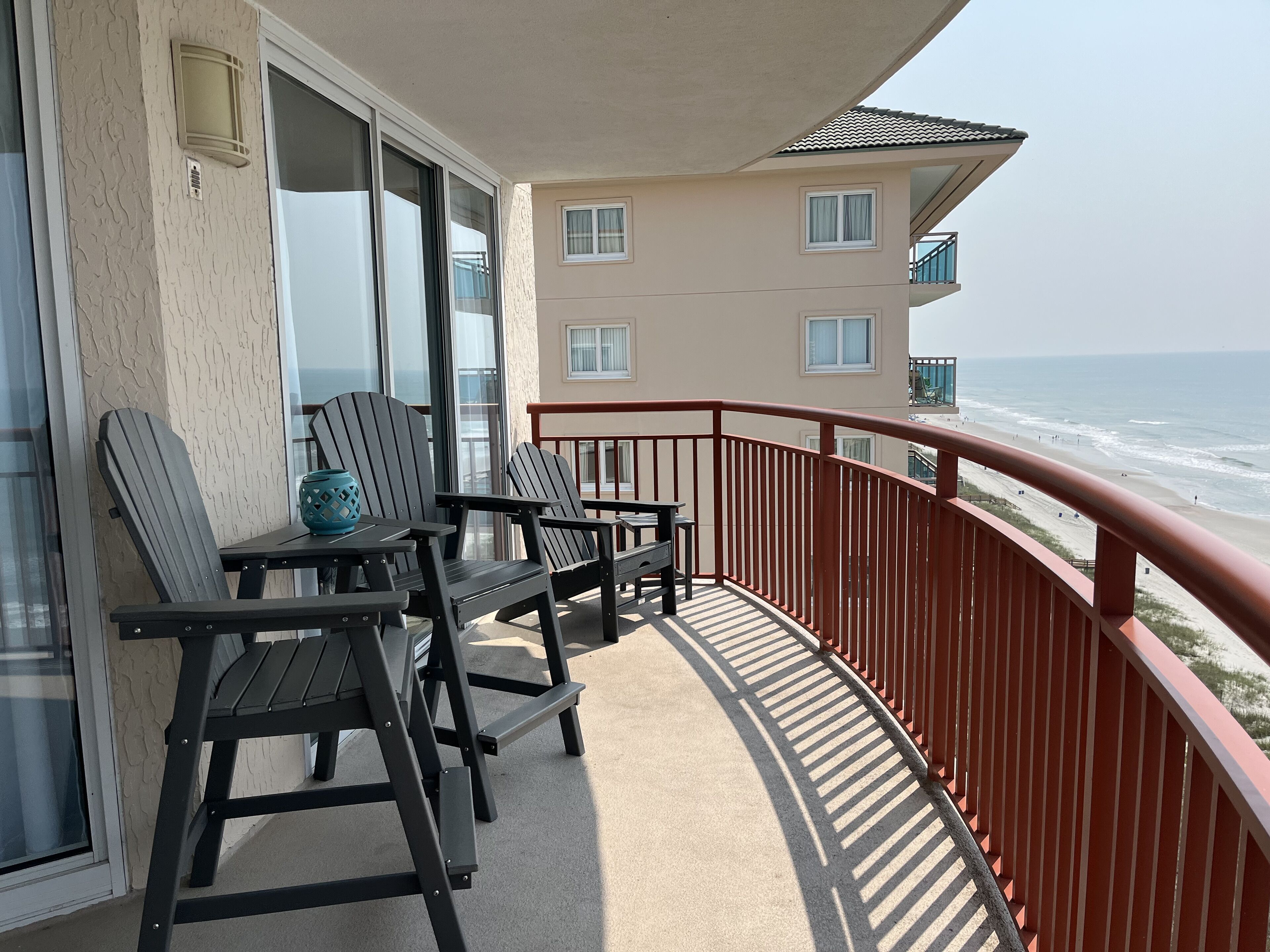 A balcony with two Adirondack chairs, a table, and a vase. The balcony overlooks a beach with a building in the background.