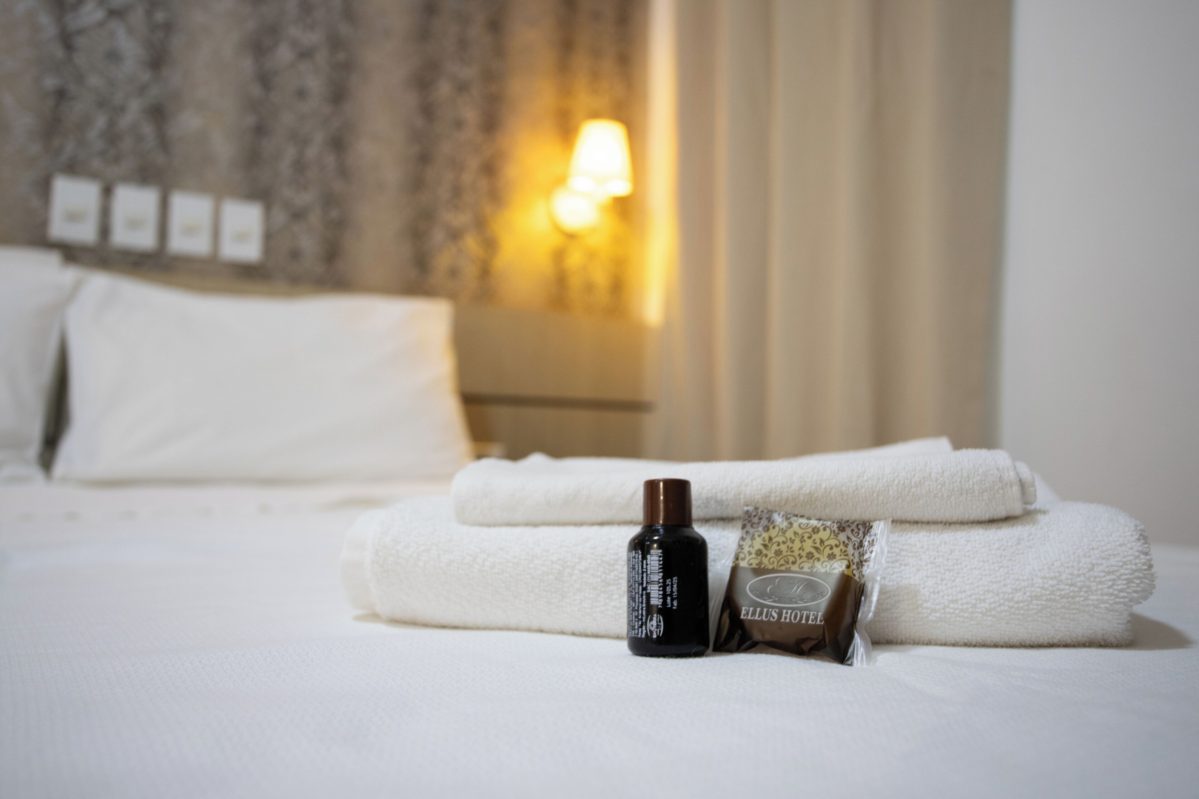 A neatly made bed with white linens, a bottle of lotion, and a towel with 'ELLUS HOTEL' branding.