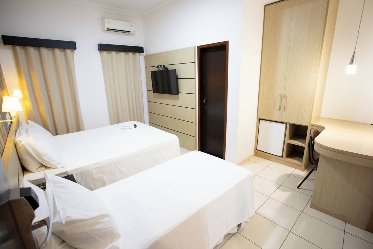 A hotel room with two beds, a TV mounted on the wall, a wardrobe, and a desk with a lamp.