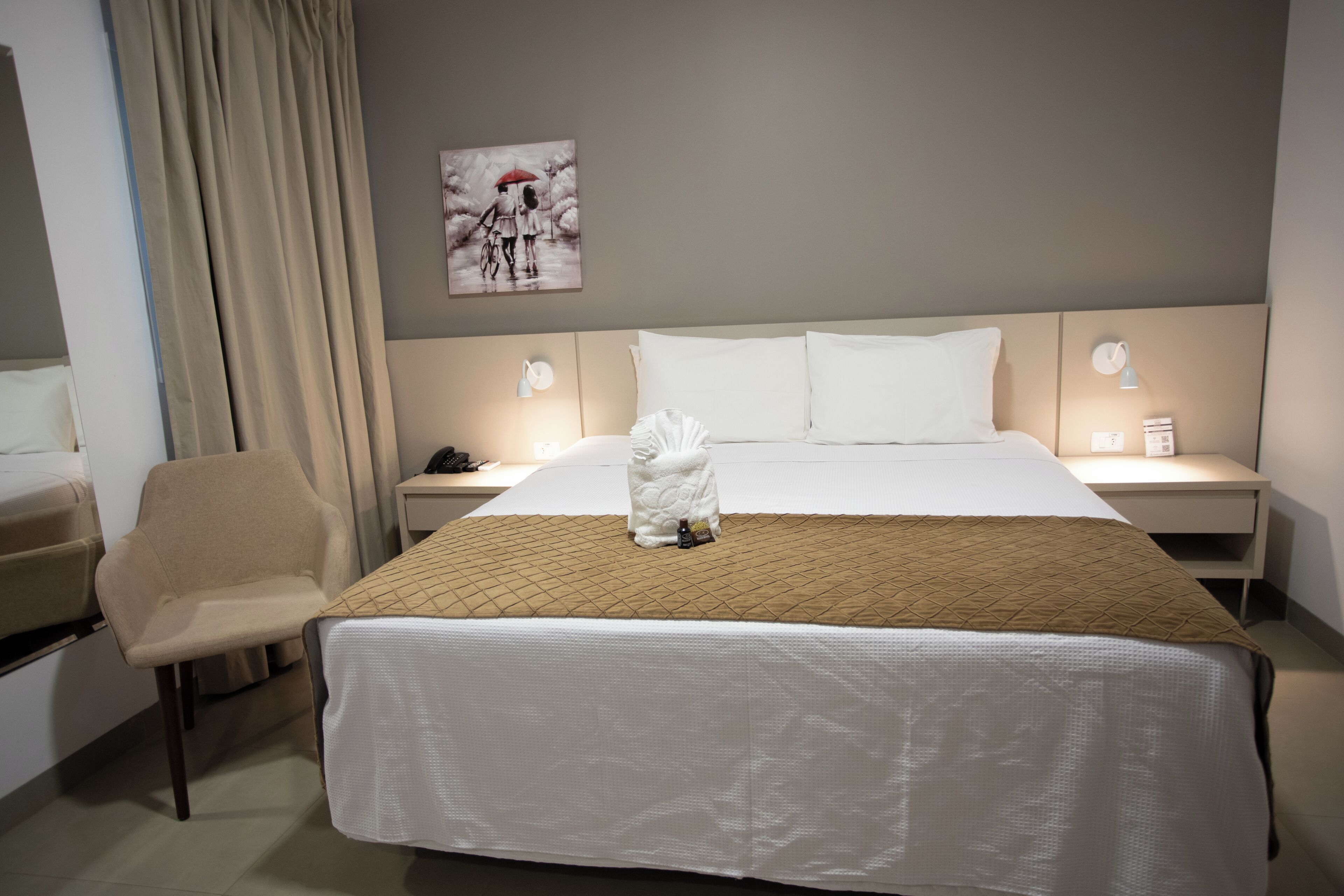 Una habitación de hotel con cama, una silla, una mesita de noche, una lámpara y un cuadro en la pared.
