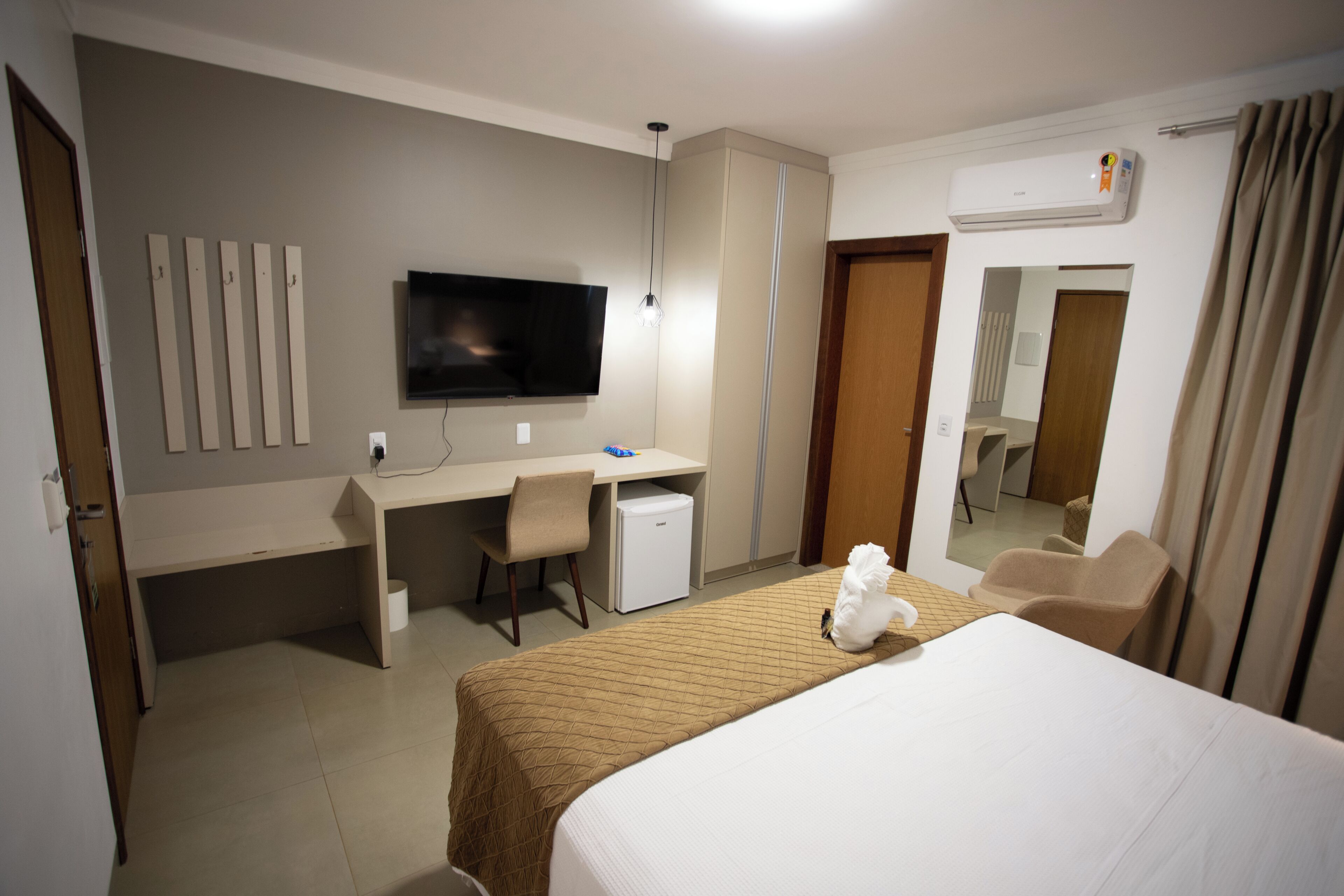 Quarto de hotel com cama, escrivaninha, cadeira, televisão e um pequeno frigorífico.
