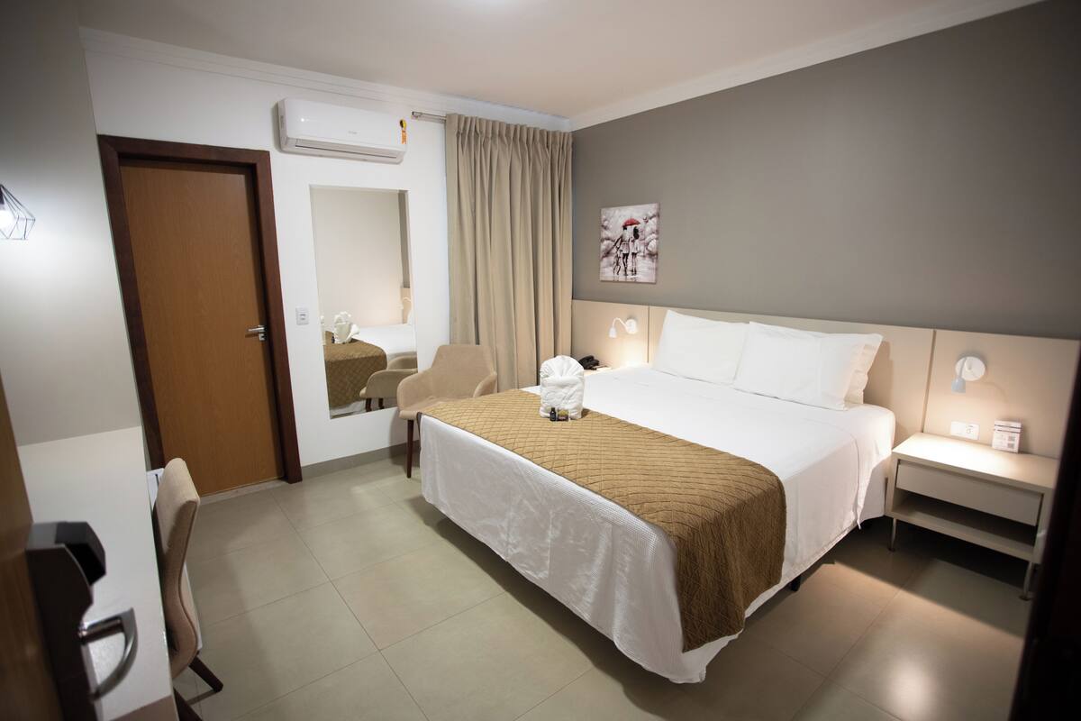 A hotel room with a bed, a chair, a desk, a nightstand, a mirror, and a picture on the wall.