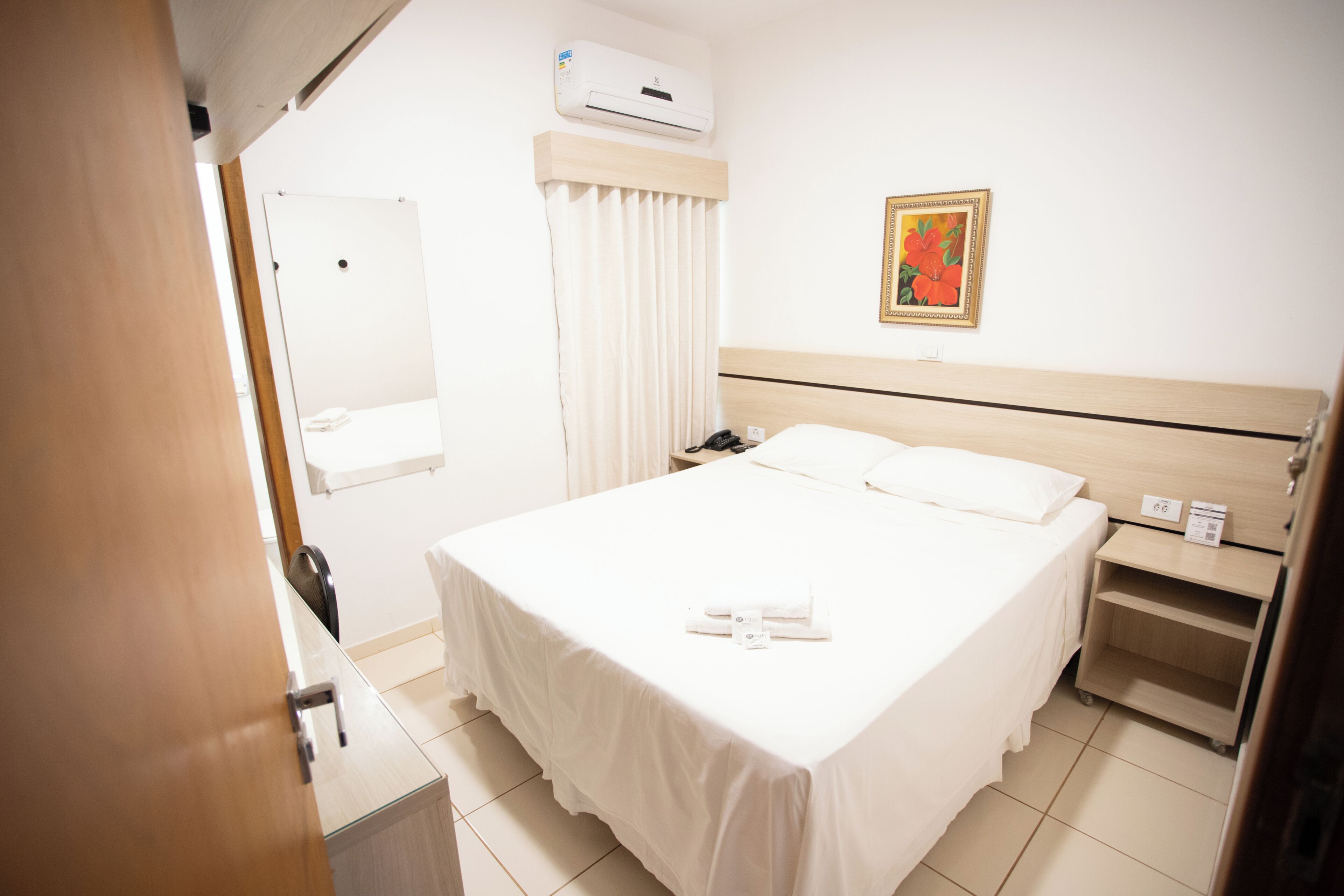 Una camera d'albergo con un letto, un comodino, uno specchio, un condizionatore d'aria e un quadro appeso al muro.