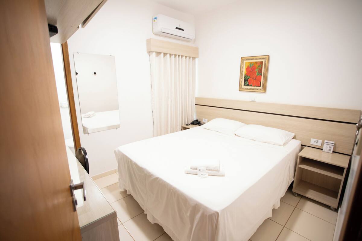 A hotel room with a bed, a bedside table, a nightstand, a mirror, a window with curtains, and an air conditioning unit.