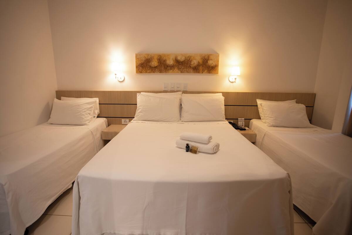 A hotel room with a large bed, two smaller beds, a headboard with a floral design, and two wall-mounted lights.