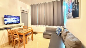 A living room with a TV, a sofa, a dining table with chairs, and a window with curtains. - Beautiful Condo -Ocean View, Best Location, Pool, Parking & Steps to All U Want (San Juan)