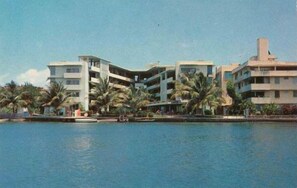 Hotel com vários andares e varandas, situado perto de um corpo d'água, com palmeiras em primeiro plano.
