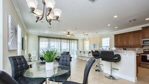 A spacious kitchen with modern appliances, a dining area with a glass table and black chairs, and a living area with a large plant and a ceiling fan. - Reunion 5 Bedroom Vacation Home 7824 (Kissimmee)