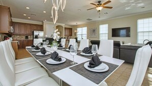A modern dining area with a long table set for a meal, a ceiling fan, and a kitchen in the background. - Reunion 5 Bedroom Vacation Home 7824 (Kissimmee)