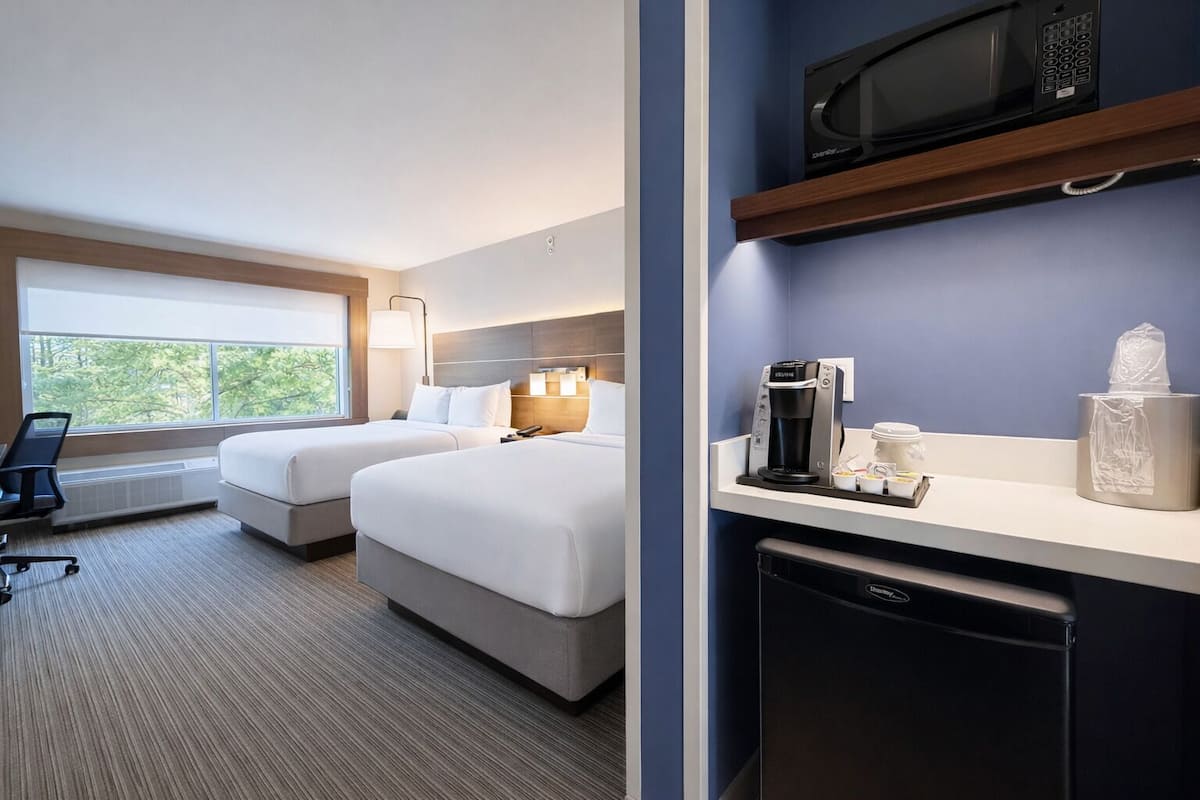 A hotel room with two beds, a desk, a computer, a microwave, a coffee maker, and a trash bin.