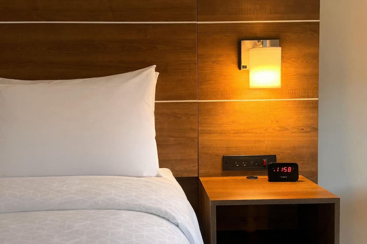 A neatly made bed with white linens and a wooden headboard. A bedside table with a digital clock and a wall-mounted light is visible.