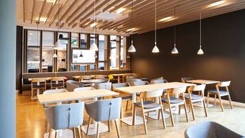 Un espace repas moderne avec des tables et des chaises en bois, des luminaires suspendus et une vue sur la cuisine en arrière-plan.