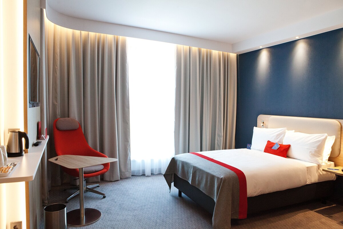 A hotel room with a bed, a desk, a chair, and a large window with curtains.