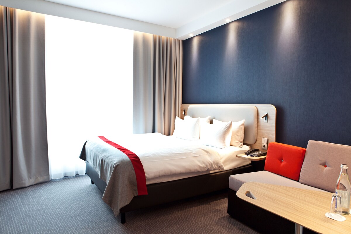 A modern hotel room with a large bed, a sofa, a small table, and a view of the outdoors.