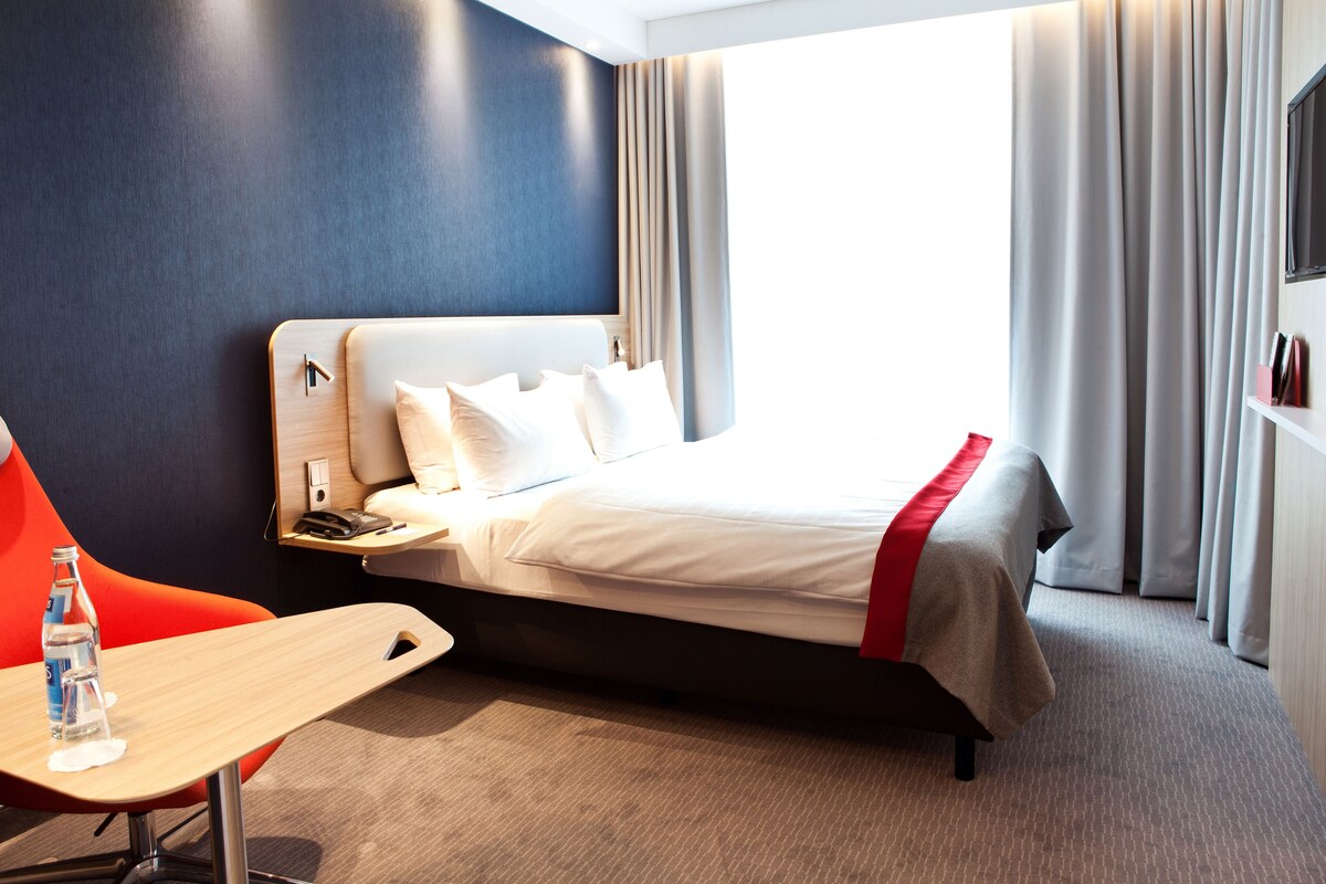 A hotel room with a bed, a desk, a chair, a water bottle, and a TV.