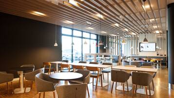 Un espace de restauration moderne avec des tables et des chaises en bois, de grandes fenêtres et des luminaires suspendus.
