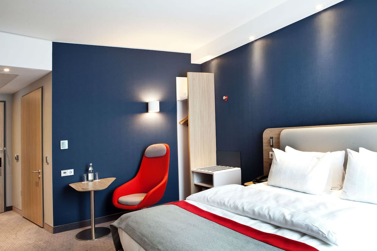 A modern hotel room with a large bed, a red chair, a small table, and a built-in wardrobe.